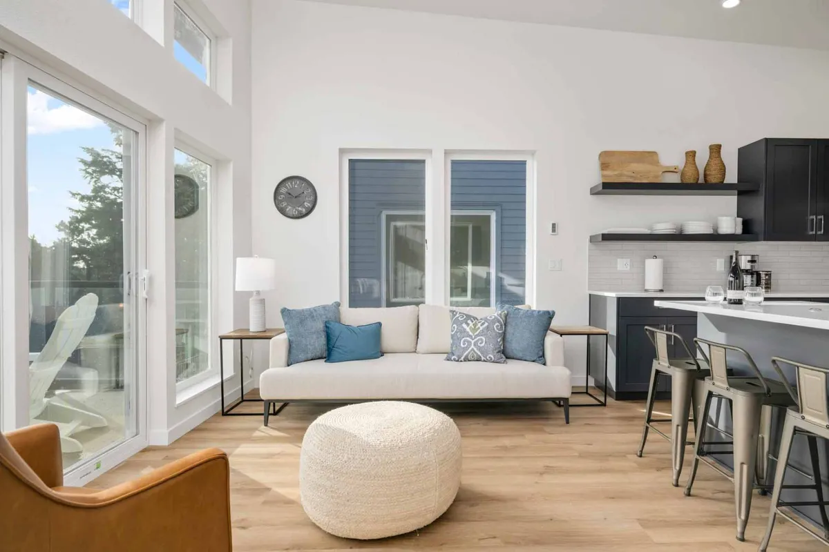 A cozy living room with plush seating, a round pouf, bar stools, and well-styled shelves. The space feels modern and welcoming, ideal for relaxing, entertaining, or enjoying casual conversations. Sunlight pours in through the windows, adding to the warmth.