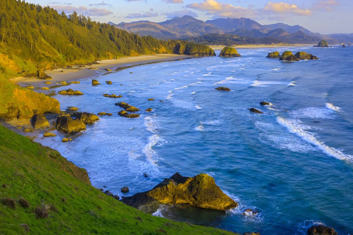 A striking coastal scene featuring rocky outcrops, foaming waves, and a sandy beach edged by a dense forest and distant mountains. The image captures the wild, natural beauty of the Oregon coast and the opportunities for exploration and discovery.
