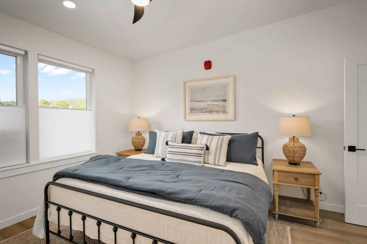 A bright and tranquil bedroom featuring a neatly made bed, soft linens, tasteful wall art, and warm table lamps. Sunlight pours in through a nearby window, creating a calm and inviting retreat perfect for restful nights.