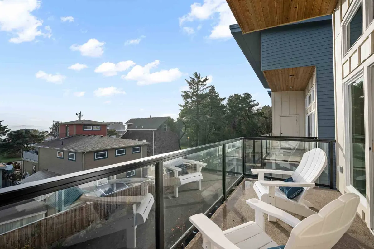 A serene balcony featuring white chairs and a view of the surrounding houses and trees. The setting is perfect for outdoor dining or relaxing, with ample natural light and a tranquil coastal atmosphere.