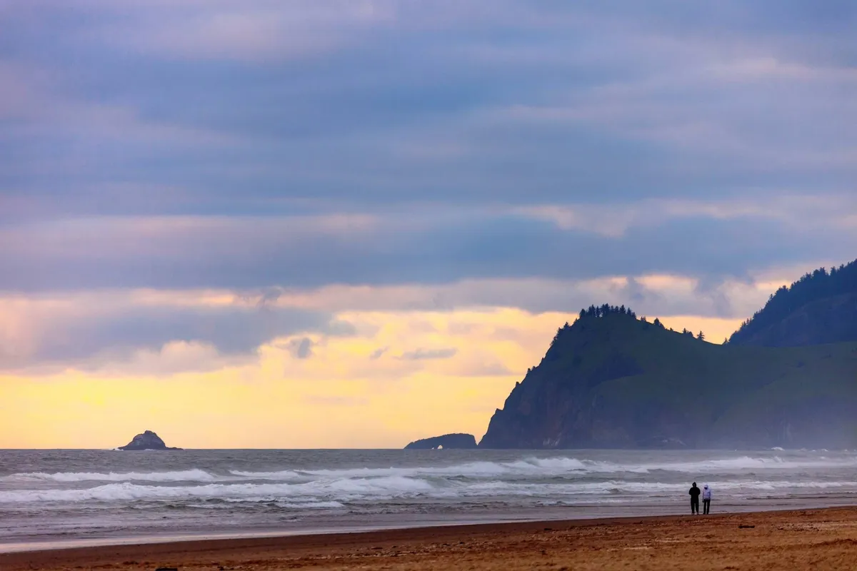 A couple walks hand-in-hand along the sandy shoreline with gentle ocean waves lapping at their feet and distant cliffs providing a scenic backdrop. The golden hour lighting suggests a peaceful, romantic sunset walk.
