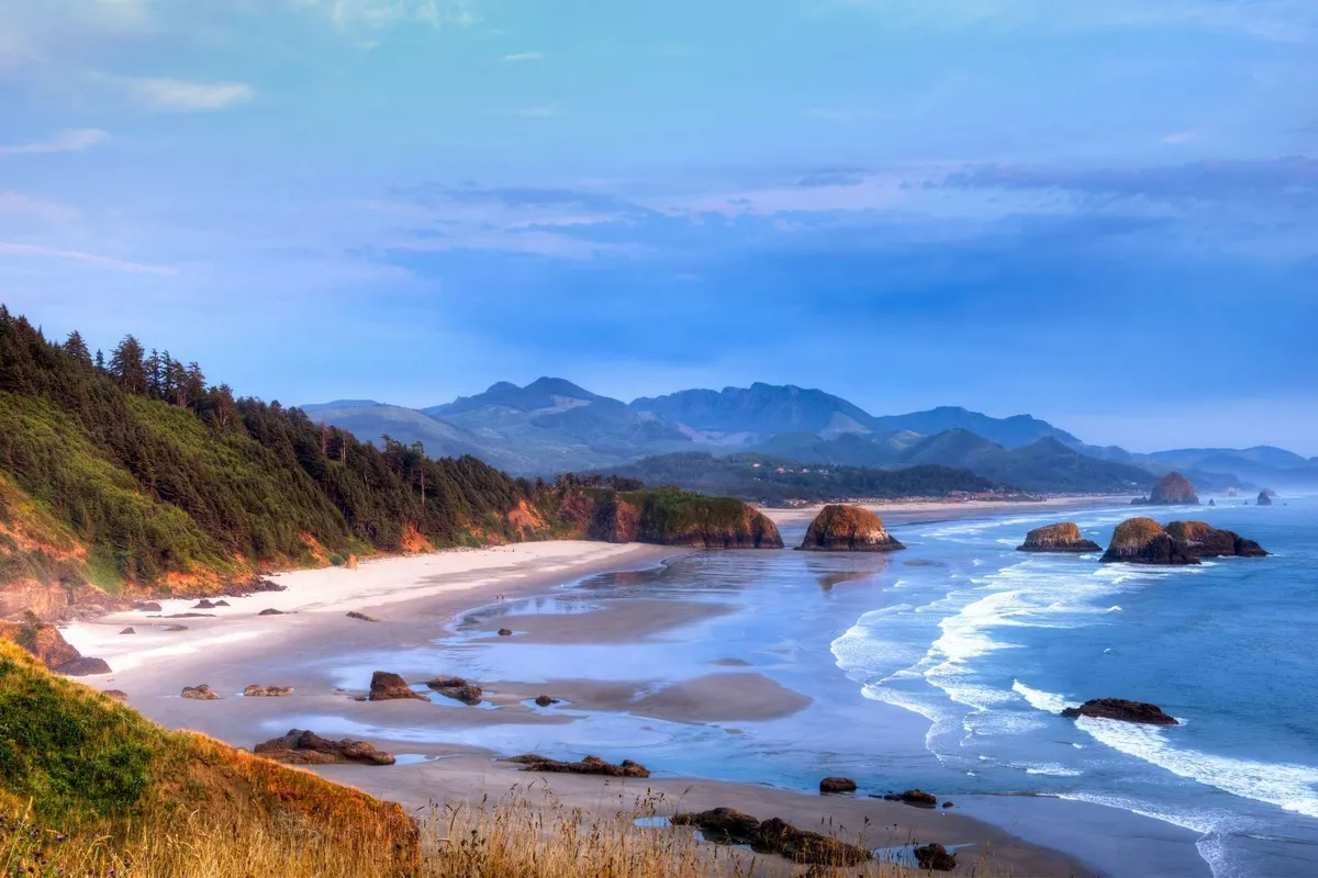 A panoramic view of Lincoln City's wide, sandy beach with the Pacific Ocean stretching out to the horizon, dramatic rock formations dotting the shoreline, and distant forested mountains framing the backdrop. The tranquil atmosphere and expanse of sand invite beachgoers to picnic, play, or simply stroll along the water's edge.