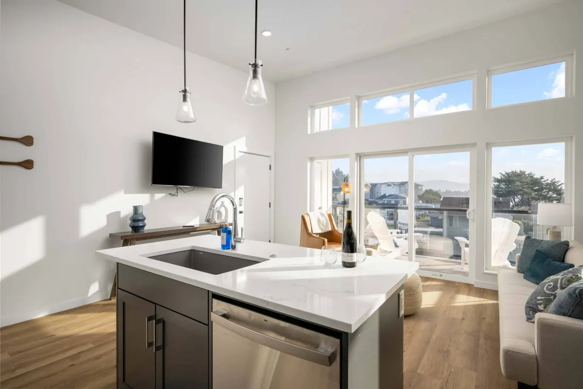 A spacious, sunlit interior showcasing an open-plan living and kitchen area. Large windows let in abundant natural light, illuminating comfortable seating, a kitchen island, and a relaxed lounge area. The layout seamlessly blends areas for relaxing and entertaining.