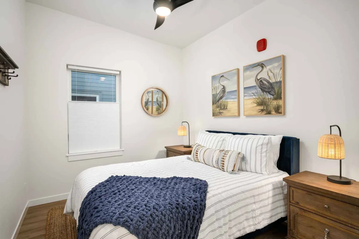 A serene bedroom scene featuring a neatly made bed with a blue blanket, matching nightstands, lamps, and tranquil artwork. Sunlight filters in, creating a peaceful environment ideal for restful nights after busy days.