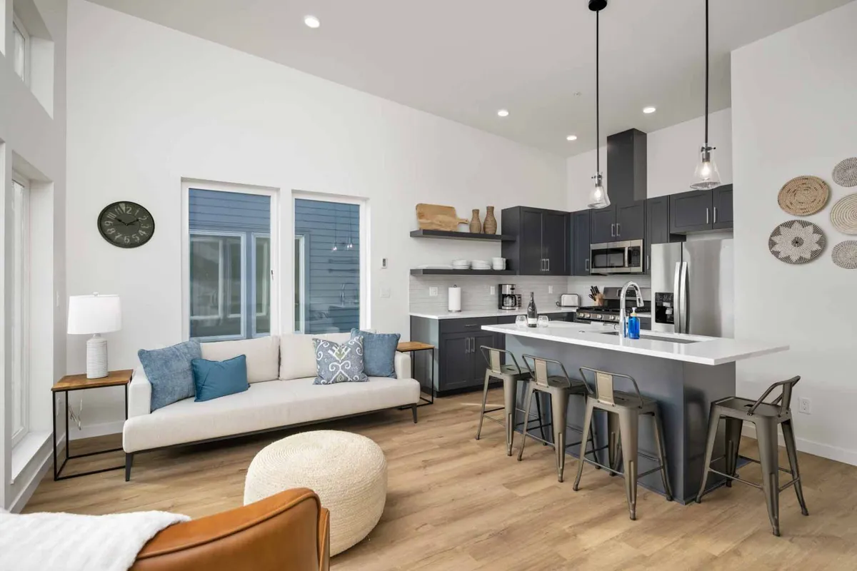 The image showcases a bright, contemporary living space featuring a cozy sofa with stylish cushions, a sleek kitchen island with bar stools, and expansive windows filling the loft with natural light. The inviting layout is perfect for relaxing, socializing, and enjoying the comforts of home within a modern coastal setting.