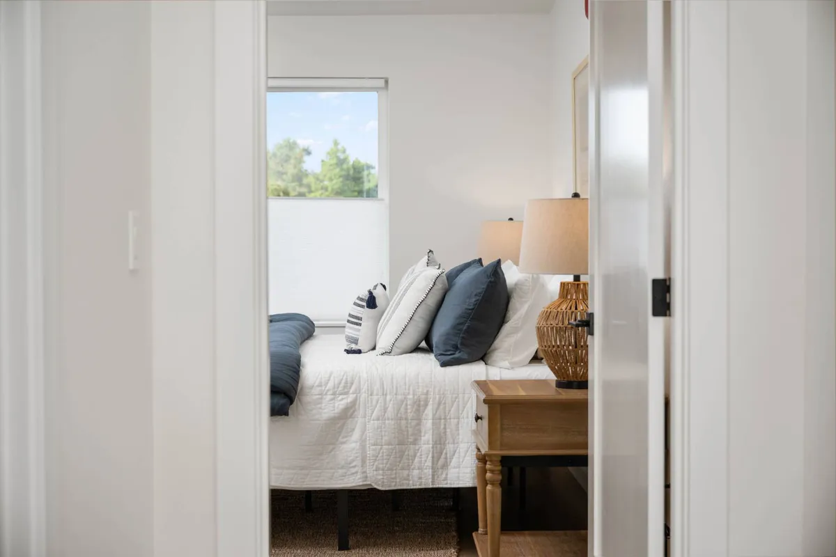 A cozy bedroom scene with a bed adorned with decorative pillows, a nightstand with a lamp, and a window offering natural daylight. The space feels calm and soothing—ideal for relaxation and privacy.